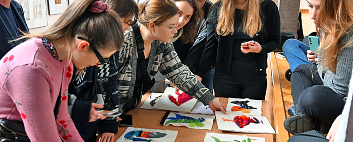 Uczestnicy warsztatów zgromadzeni przy stole, na którym rozłożone zostały wykonane przez nich ilustracje. Prowadząca warsztaty omawia prace